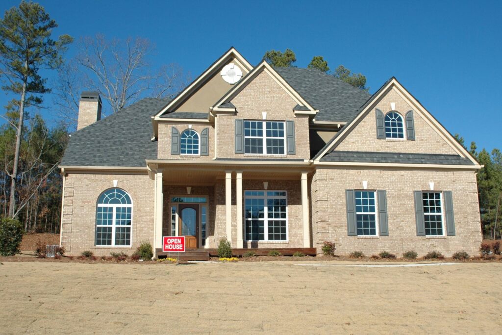 A large, brown, two-story brick home sits in a large yard on a sunny day with a red open house sign planted in front.