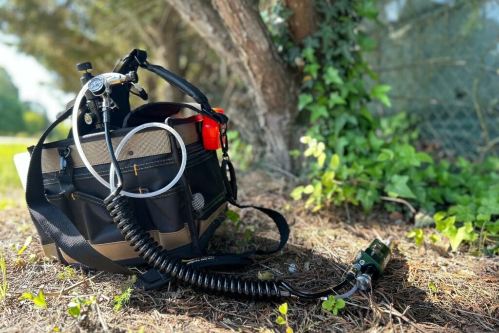 A large tool bag filled with tree healthcare tools, including a tree injection system, is lying on the ground next to some trees.