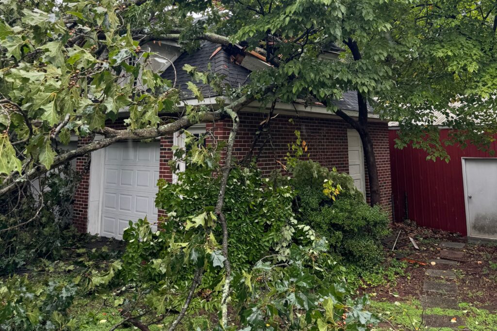 Large fallen tree leaning against a brick garage with severe roof damage and scattered debris across the driveway after high winds.