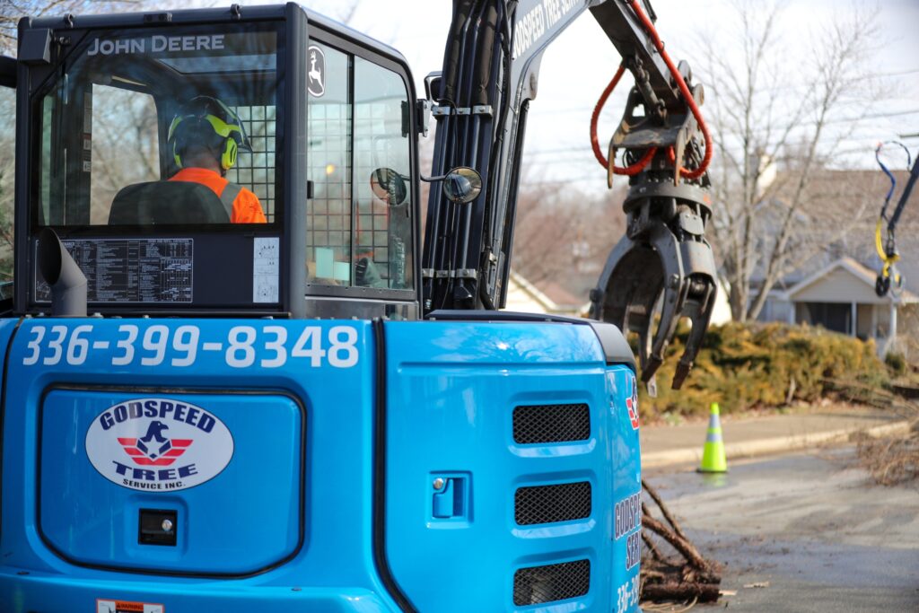 Godspeed Tree Service operator in a blue John Deere skid steer uses a grapple attachment to remove storm debris in a Winston-Salem neighborhood.