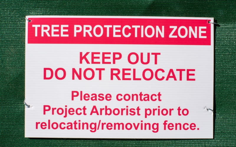 Close-up of a tree protection zone sign on a construction site in Winston-Salem. The sign has bold red lettering that reads “Tree Protection Zone –Keep Out, Do Not Relocate, please contact project arborist prior to relocating/removing fence.”