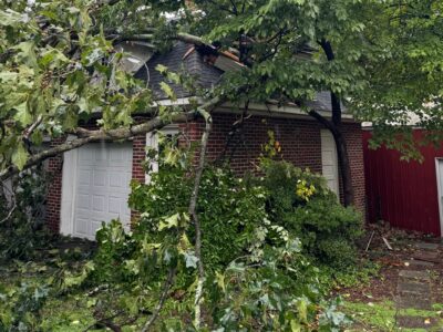 Large fallen tree leaning against a brick garage with severe roof damage and scattered debris across the driveway after high winds.